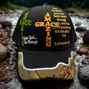 Op de afbeelding: Zwarte baseballcap met "AMAZING GRACE" geborduurd in goud en groen. De cap heeft de tekst "Seek God... See Change" en een schriftgedeelte uit Efeziërs 2:8. De klep heeft een camouflagepatroon.