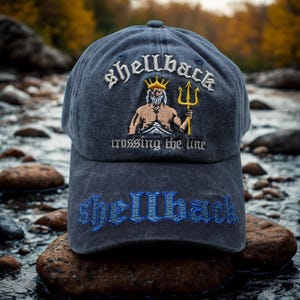 Peut inclure: Casquette de baseball bleu marine avec le mot "shellback" brodé en blanc au-dessus d'une illustration d'une figure musclée avec un trident. L'expression "crossing the line" est en dessous, et "shellback" est brodé en bleu sur la visière.