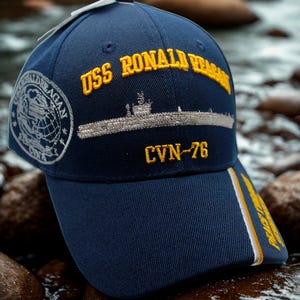 Op de afbeelding: Marineblauwe baseballcap met gouden borduursel "USS Ronald Reagan". De cap heeft een geborduurd beeld van een onderzee&euml;r en "CVN-76". Een zijpatch toont "Ronald Reagan" en een wereldbol.