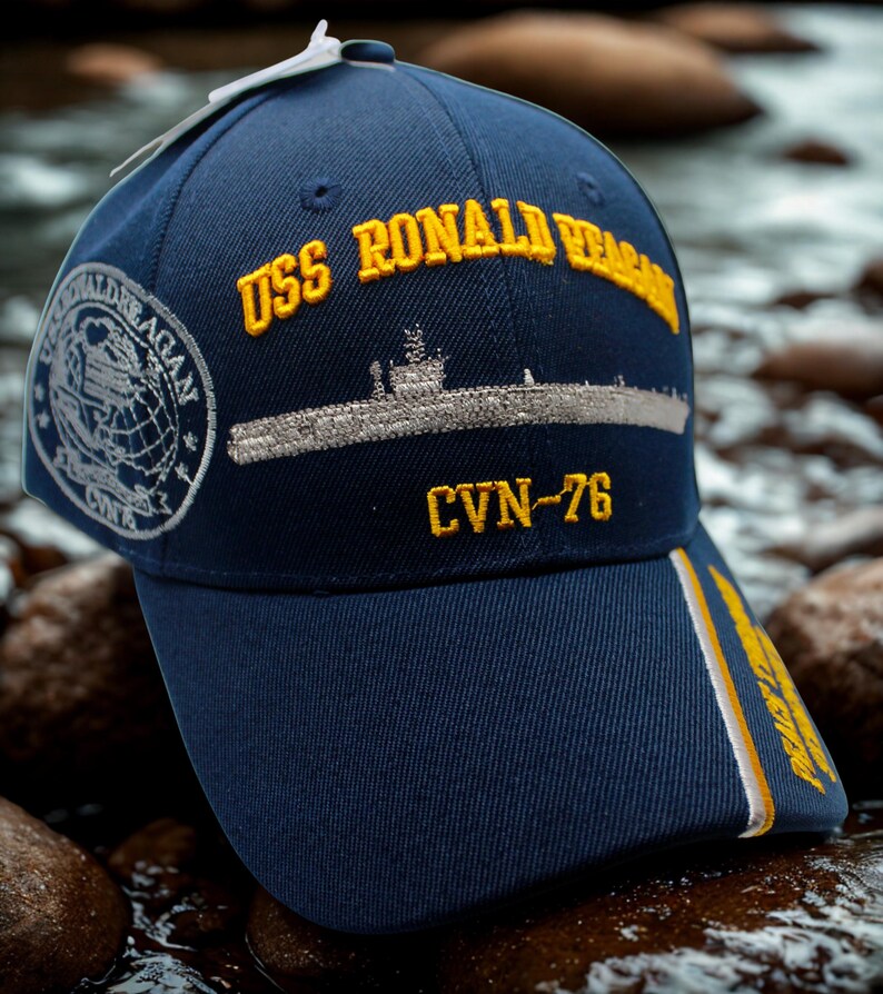 Op de afbeelding: Marineblauwe baseballcap met gouden borduursel "USS Ronald Reagan". De cap heeft een geborduurd beeld van een onderzee&euml;r en "CVN-76". Een zijpatch toont "Ronald Reagan" en een wereldbol.