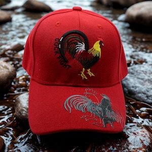 May include: Red baseball cap with embroidered rooster design. The cap features the text "COCK FIGHT" and a detailed image of a rooster on the front. The brim displays a second rooster design.