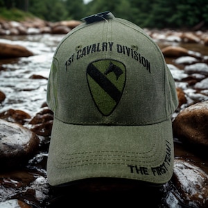 May include: Olive green baseball cap with the text "1st Cavalry Division" and a shield emblem. The emblem features a black diagonal stripe and a green background. The text "THE FIRST TEAM!" is embroidered on the brim.