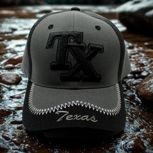 May include: A grey and black baseball cap featuring the letters "TX" in black on the front. The black brim has "Texas" embroidered in white. The cap has a curved peak and a structured crown.