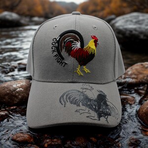 May include: A grey baseball cap with an embroidered rooster design. The cap has the text "COCK FIGHT" above the rooster. The rooster is in red, yellow, and black. A second rooster design is on the brim. The cap is a classic style.
