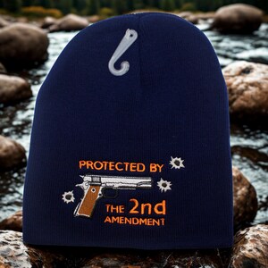 May include: Navy blue beanie with a white plastic hang tag. The beanie features an embroidered design of a handgun, bullet holes, and the text "PROTECTED BY THE 2nd AMENDMENT" in orange.