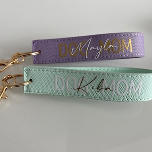 May include: Two purple and light green keychains with gold key rings. The keychains are made of fabric and have the text "Dog Mom" written on them. The purple keychain has the name "Mayla" written in gold lettering and the light green keychain has the name "Kiba" written in gold lettering.