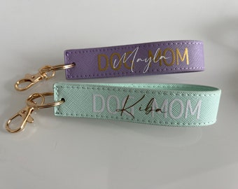 May include: Two purple and light green keychains with gold key rings. The keychains are made of fabric and have the text "Dog Mom" written on them. The purple keychain has the name "Mayla" written in gold lettering and the light green keychain has the name "Kiba" written in gold lettering.