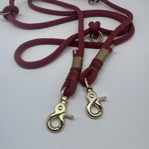 K&ouml;nnte beinhalten: Eine rote Hundeleine aus Seil mit zwei goldenen Metallclips und zwei goldenen Metallringen. Die Leine ist aus geflochtenem Seil und hat einen braunen Akzent.