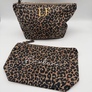 Könnte beinhalten: Zwei Kosmetiktaschen mit Leopardenmuster. Die größere Tasche hat goldfarbene Initialen "LF" und einen Reißverschluss. Die kleinere Tasche trägt den Namen "Barbara" in weißer Schrift. Beide Taschen haben ein braun-schwarzes Fleckenmuster.