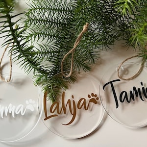 Könnte beinhalten: Drei runde, transparente Acryl-Weihnachtsornamente mit Namen und Pfotenabdrücken. Die Namen "Emma" in Weiß, "Lahja" in Gold und "Tami" in Schwarz sind abgebildet. Jedes Ornament hat eine Schlaufe aus Jute zum Aufhängen, vor grünem Laub.