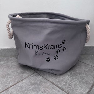 Könnte beinhalten: Grauer Canvas-Aufbewahrungskorb mit Seilgriffen. Der Korb trägt den Text "KrimsKrams Kiba" und hat bedruckte Pfotenabdrücke.