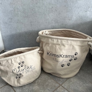 Könnte beinhalten: Zwei beigefarbene Aufbewahrungskörbe aus Canvas mit schwarzen Pfotenabdrücken und Text. Der größere Korb trägt die Aufschrift "Krimskrams Mayla" und der kleinere Korb die Aufschrift "Maylas Spielzeug".
