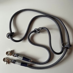 May include: A gray double dog leash with two silver clips and a silver ring. The leash is made of rope and has a navy blue and white stripe on each clip.