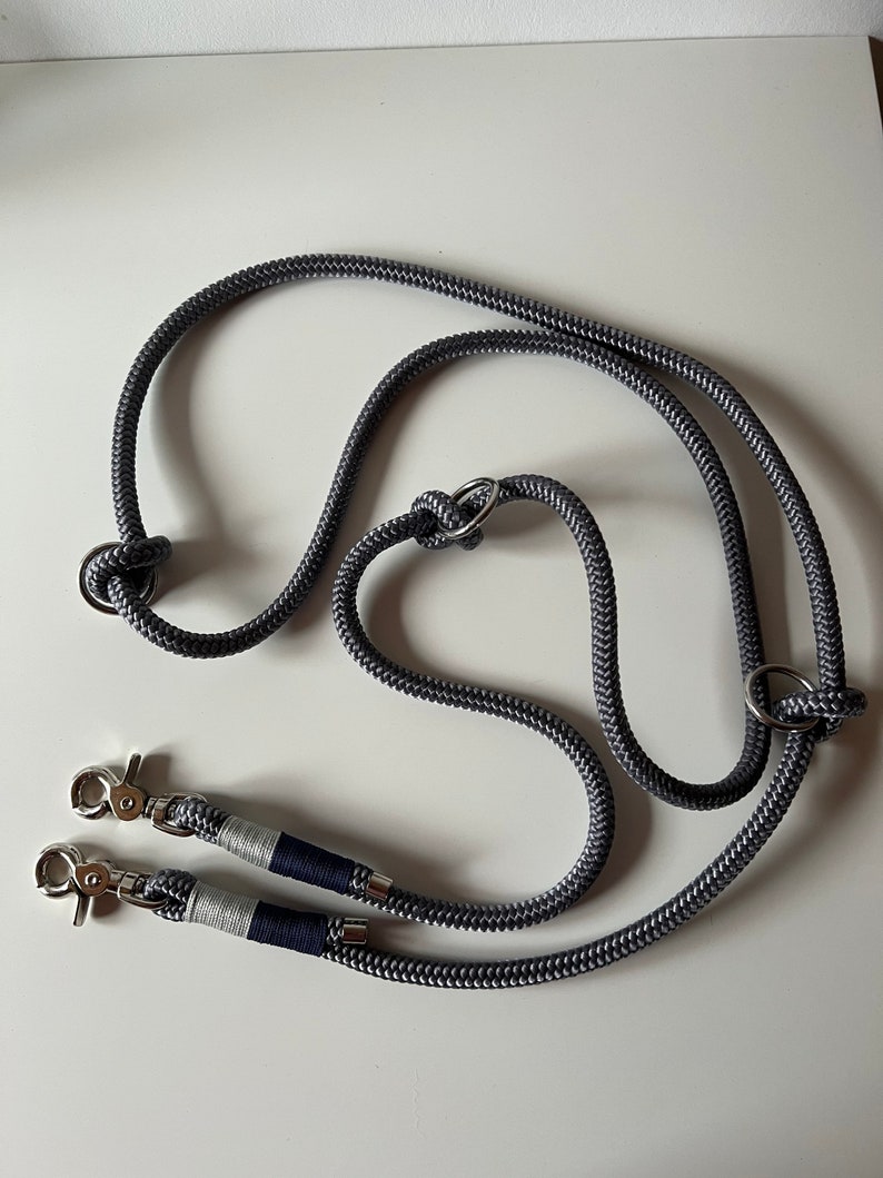 May include: A gray double dog leash with two silver clips and a silver ring. The leash is made of rope and has a navy blue and white stripe on each clip.