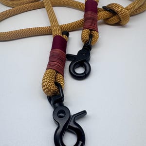K&ouml;nnte beinhalten: Eine goldene Hundeleine aus Seil mit einem burgunderroten und roten Akzent und zwei schwarzen Metallclips.