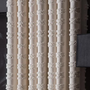 May include: An off-white curtain panel featuring vertical rows of raised, tufted texture. Beside the curtain stands a black table lamp with a rectangular shade, positioned next to two white ceramic jars with gold-toned lids on a metal table.