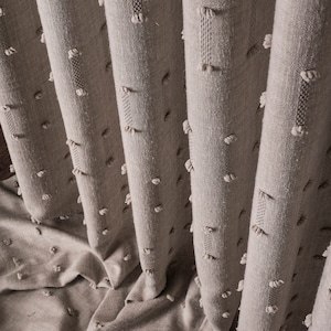 May include: Close-up of a beige textured fabric curtain with small white pom-poms. The fabric has a subtle woven pattern.