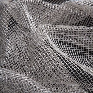May include: Close-up of a silver metallic mesh fabric with a square grid pattern. The fabric is draped, creating folds and shadows. The material has a shiny, reflective quality, and the background is black.