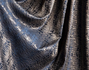 May include: Close-up of a draped fabric with a textured pattern. The fabric has a dark blue base with a shimmering, mottled design in shades of silver and grey. The folds create shadows and highlights.