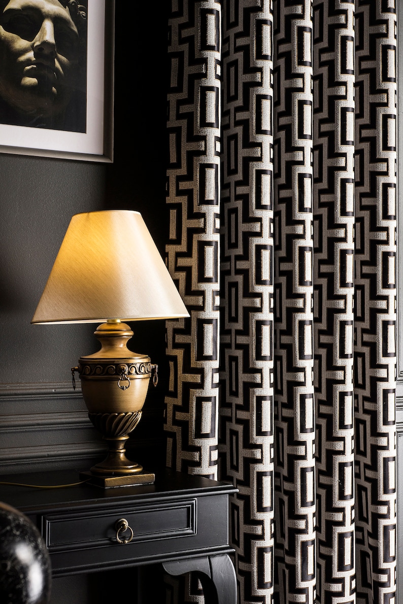 May include: A gold table lamp with a white lampshade sits on a black side table with a drawer. The lamp is in front of a black and white geometric patterned curtain.