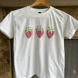 May include: White t-shirt with three red and white gingham strawberries appliqued on the front.
