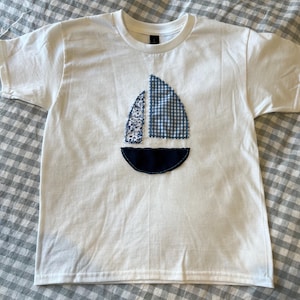 May include: A white t-shirt with a blue and white sailboat design sewn on the front. The sailboat is made of two different fabrics, one with a blue and white gingham pattern and the other with a white floral pattern.