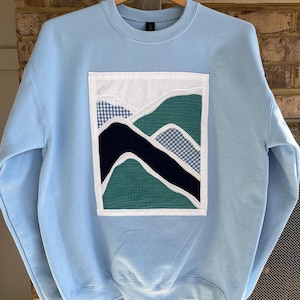 May include: A light blue sweatshirt with a rectangular patch featuring a mountain landscape design. The patch includes white, green, navy, and gingham patterned fabric, creating a layered effect. The sweatshirt is hanging on a wooden hanger.
