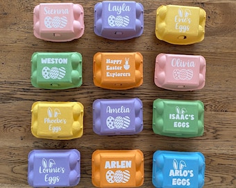 Peut inclure: Plusieurs boîtes à œufs de Pâques colorées, chacune avec un nom ou un message imprimé sur le couvercle. Les boîtes sont de différentes couleurs pastel, dont rose, violet, vert, jaune, orange et bleu. Elles sont disposées sur une surface en bois.