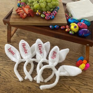 May include: A wooden serving tray with grapes, strawberries, and chocolate eggs. White bunny ear headbands with names embroidered in red are in the foreground. A colourful Easter-themed arrangement.