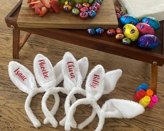 May include: A wooden serving tray with grapes, strawberries, and chocolate eggs. White bunny ear headbands with names embroidered in red are in the foreground. A colorful Easter-themed arrangement.