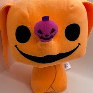 Peut inclure: Une peluche avec une grande tête carrée orange, des yeux noirs circulaires et un large sourire noir fin. Un petit nez violet en forme de citrouille d'Halloween avec une tige violet foncé et un visage noir sculpté est situé au centre du visage.