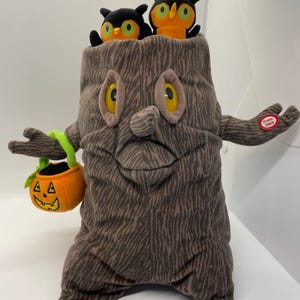 Könnte beinhalten: Ein Halloween-Dekoration aus Plüsch in Form eines Baumstumpfes mit einem lächelnden Gesicht, großen gelben Augen und zwei Plüsch-Eulen, die aus der Oberseite herausragen. Es hat ausgestreckte Arme, einer hält einen kürbisförmigen Eimer. Der andere Arm hat einen "Press Here"-Knopf.