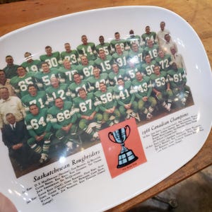 May include: A white ceramic plate featuring a photograph of the 1966 Saskatchewan Roughriders football team. The team is wearing green and white uniforms and holding a trophy. The text "Saskatchewan Roughriders" and "1966 Canadian Champions" is printed on the plate.