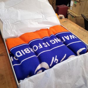 Può includere: Un maglione di lana blu e arancione con il testo "WAITING IS FORBIDDEN" in lettere bianche. Il maglione è piegato e posto all'interno di una scatola foderata con carta velina bianca.