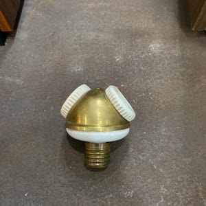May include: A brass-colored bottle stopper with a white base and three white, ridged accents. The stopper has a threaded base and a conical top. The object is designed for use with bottles.