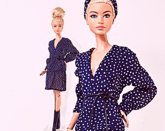 May include: Two fashion dolls wearing matching navy blue dresses with white polka dots. The dresses feature long sleeves, a V-neckline, and a thin black tie belt at the waist. One doll has a high bun hairstyle, while the other wears a matching headband.