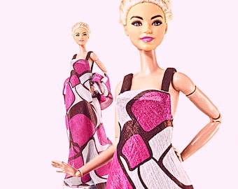 Op de afbeelding: Twee modepoppen in lange jurken met een roze, witte en bruine geometrische print. De poppen hebben blond haar en staan tegen een lichtroze achtergrond. De jurken hebben donkerbruine bandjes.