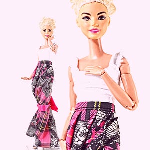 May include: Two dolls with blonde hair styled in short, textured cuts. Each doll wears a white sleeveless top with ruffled straps and a long, pleated skirt featuring a black, white, and pink plaid pattern with additional graphic prints.
