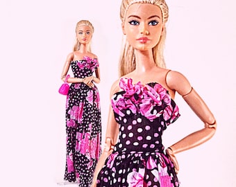 Op de afbeelding: Twee poppen met blond haar in een paardenstaart, gekleed in strapless, vloerlange jurken van zwarte stof met witte stippen en roze bloemenpatronen. De poppen hebben beweegbare armen en een van hen houdt een klein, felroze handtasje vast.