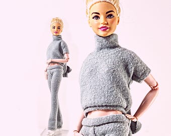 May include: A doll with blonde hair styled in an updo, wearing a grey short-sleeved turtleneck sweater and matching grey pants. The doll has articulated joints at the elbows and wrists, and is shown in two positions, one close-up and one full-body.