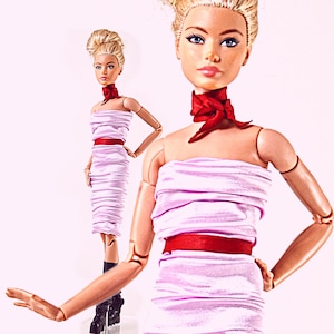 May include: A blonde doll with articulated joints wearing a strapless, light pink ruched dress with a red satin waistband and a matching red neck scarf. The doll has painted facial features and is styled with an updo hairstyle.