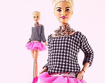 Op de afbeelding: Een modepop met een zwart-wit patroon trui, een roze tutu rok en roze laarzen. De pop heeft een zilveren ketting en een riem. De pop is blond met roze lippenstift.