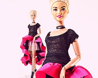 Op de afbeelding: Een modepop met een zwarte kanten top en een felroze en zwarte rok. De pop heeft blond haar en een zwarte choker. De rok heeft een gelaagd ontwerp met een roze buitenlaag en een lichtere roze onderlaag. De pop draagt zwarte laarzen.