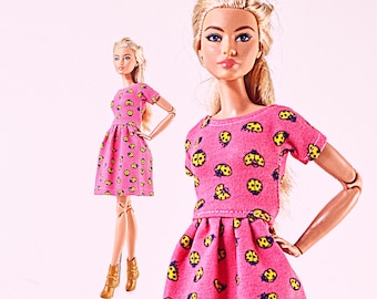 Op de afbeelding: Pop in een roze jurk met een geel lieveheersbeestjespatroon. De pop heeft blond haar en gouden laarzen. De jurk heeft korte mouwen en een getailleerde taille. De achtergrond is zachtroze.