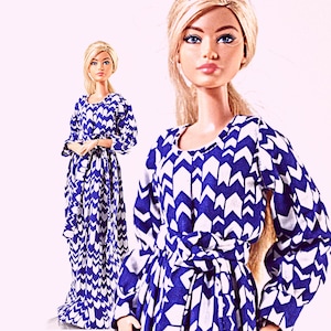 May include: Two dolls with blonde hair styled in a ponytail, each wearing a long-sleeved, floor-length dress featuring a repeating blue and white chevron pattern. The dresses have a round neckline and a matching fabric tie belt at the waist.