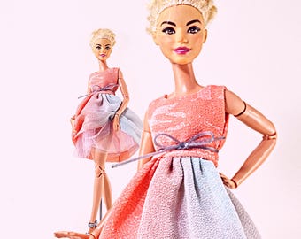 Op de afbeelding: Twee poppen met blond haar in een opgestoken kapsel, gekleed in mouwloze jurken met een gestructureerd koraalroze lijfje en een plooirok met afwisselende panelen van koraalroze en lichtblauwe stof, geaccentueerd door een dun paars lint in de taille.