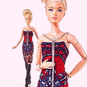 May include: Two dolls with blonde hair styled in high buns, wearing strapless red, black, and white patterned dresses. Each doll features a silver-toned chain necklace that drapes down the centre of the dress. The dolls have articulated arms and are wearing black boots.