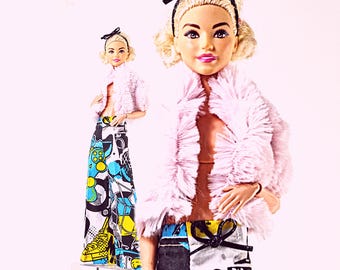 May include: A doll with blonde hair styled with a black headband, wearing a light pink faux fur cropped jacket over a tan top. The doll wears wide-leg trousers featuring a graphic print in black, white, yellow, and blue, with a black bow at the waist.