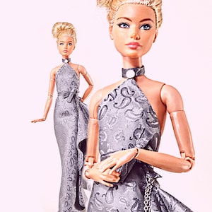 May include: Two articulated dolls with blonde hair styled in an updo, wearing floor-length silver-grey dresses with a dark paisley pattern. The dresses feature a halter neckline with a white circular accent at the neck and a side ruffle detail.