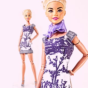 Puede incluir: Una muñeca con un vestido blanco y morado con un diseño de árbol. El vestido tiene un dobladillo con flecos y mangas cortas. La muñeca también lleva una bufanda morada y tacones plateados. La muñeca tiene el pelo rubio.